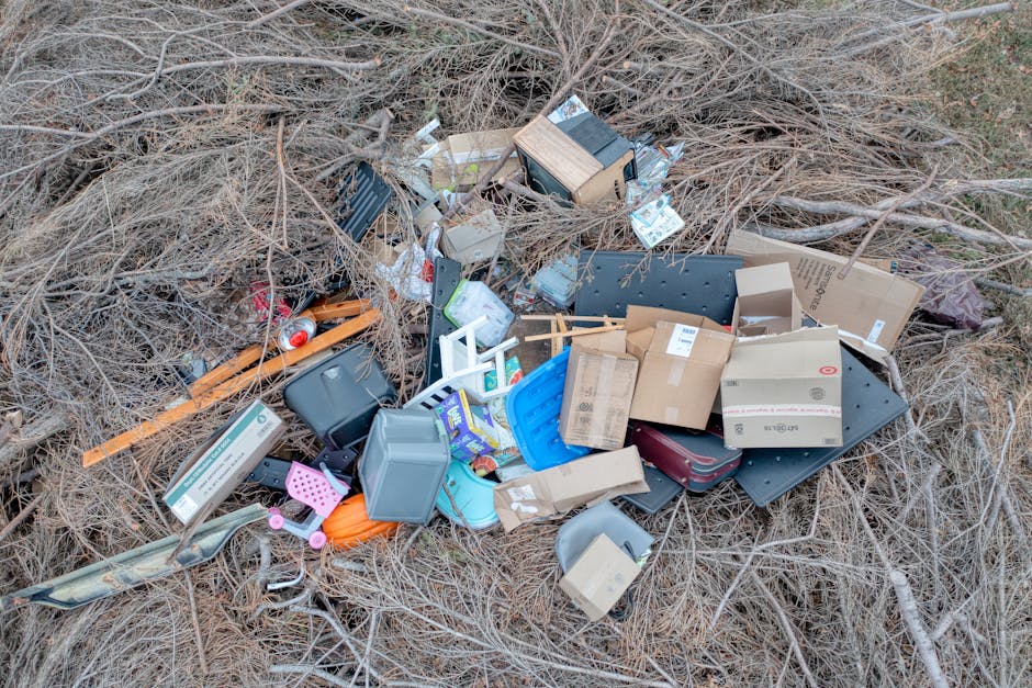 A large pile of discarded household waste and debris, including cardboard boxes, plastic containers, packaging materials, and miscellaneous household items, lies on a patch of dry grass and leafless bushes outdoors. The rubbish appears to be a mixture of broken furniture parts, packaging, and waste materials, reflecting improper disposal. The scene is set on the ground outside a property, with no visible vehicles or equipment. The image suggests the context of waste clearance, which could relate to house removals or property clearance services provided by Man with Van Millwall, as referenced on the webpage about avoiding fines for bulky waste disposal in Millwall (Tower Hamlets). The natural lighting highlights the cluttered and unmanaged state of the waste, emphasizing the importance of professional disposal and relocation services.