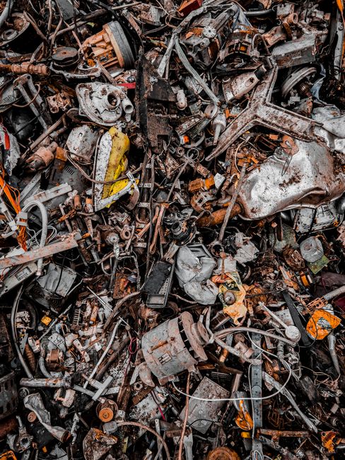 A large pile of assorted scrap metal and vehicle parts, including engine components, bolts, wires, and panels, all tangled together in a dense heap. The metal pieces are rusted and weathered, with some painted surfaces peeling or corroded. The collection is situated outdoors on a paved surface, with no clear boundaries or containers visible. The image captures the chaotic arrangement typical of dismantled automotive parts awaiting recycling or disposal, reflecting the type of bulky waste that might be relevant to services like those offered by Man with Van Millwall for waste removal or furniture transport during home relocation or decluttering processes.