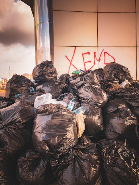 A large pile of discarded household waste and debris, including cardboard boxes, plastic containers, packaging materials, and miscellaneous household items, lies on a patch of dry grass and leafless bushes outdoors. The rubbish appears to be a mixture of broken furniture parts, packaging, and waste materials, reflecting improper disposal. The scene is set on the ground outside a property, with no visible vehicles or equipment. The image suggests the context of waste clearance, which could relate to house removals or property clearance services provided by Man with Van Millwall, as referenced on the webpage about avoiding fines for bulky waste disposal in Millwall (Tower Hamlets). The natural lighting highlights the cluttered and unmanaged state of the waste, emphasizing the importance of professional disposal and relocation services.