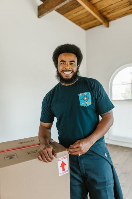 A smiling man with an afro hairstyle, dressed in a dark teal t-shirt with a patterned pocket, is indoors in a room with white walls and a wooden ceiling. He is holding and steadying a large cardboard box with a red 'CAUTION' arrow sticker, indicating careful handling during a home relocation. The room features a rounded window allowing natural light to enter. This scene depicts a professional moving process, with the man representing a mover from Man with Van Millwall as part of their furniture transport and packing services. In the background, the floor appears wooden, and the environment suggests an organized, clean space engaged in household packing and loading activities for house removals, consistent with professional moving services for flats or estate properties like Mudchute Estate in Millwall.