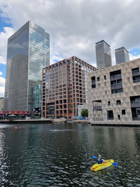 Image of a cityscape showing modern high-rise buildings overlooking a river, with a cloudy sky overhead. In the foreground, a person is lying on a yellow kayak, paddling on the water. The waterfront area features a paved walkway and a low barrier alongside the river. The buildings include a tall glass-fronted skyscraper, a reddish-brown office block with a grid of windows, and a white structure with geometric patterns. This urban scene captures the skyline near Millwall, with no visible interior or moving equipment related to house removals. The image reflects the environment surrounding the area where Man with Van Millwall offers house removals and moving services, though no specific items or furniture are depicted.