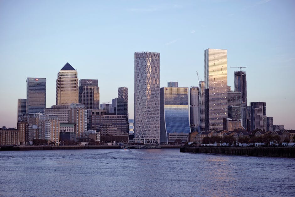 A panoramic view of the Canary Wharf skyline taken from across the Thames River during daylight hours, featuring numerous modern glass and steel high-rise office buildings, including prominent structures like the One Canada Square tower with its pyramid-shaped top. The scene shows the waterfront with calm waters reflecting parts of the cityscape, and some cranes are visible on the horizon indicating ongoing construction. The lighting suggests late afternoon or early evening, with a clear blue sky overhead. This urban landscape highlights the area's commercial prominence and contemporary architecture. The image aligns with themes of home relocation, furniture transport, and moving logistics, as seen in professional removals services such as those offered by Man with Van Millwall, who facilitate house and office moves in the Millwall and Canary Wharf area.