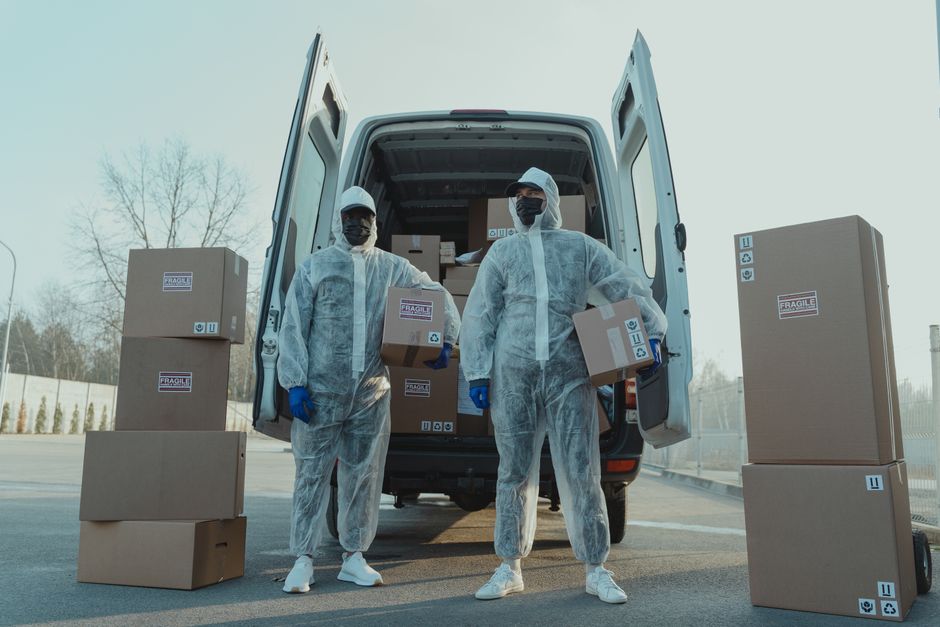 Two professional movers from Man with Van Millwall dressed in white protective suits, face masks, and blue gloves are loading cardboard boxes into the rear of a white moving van parked outdoors on a p