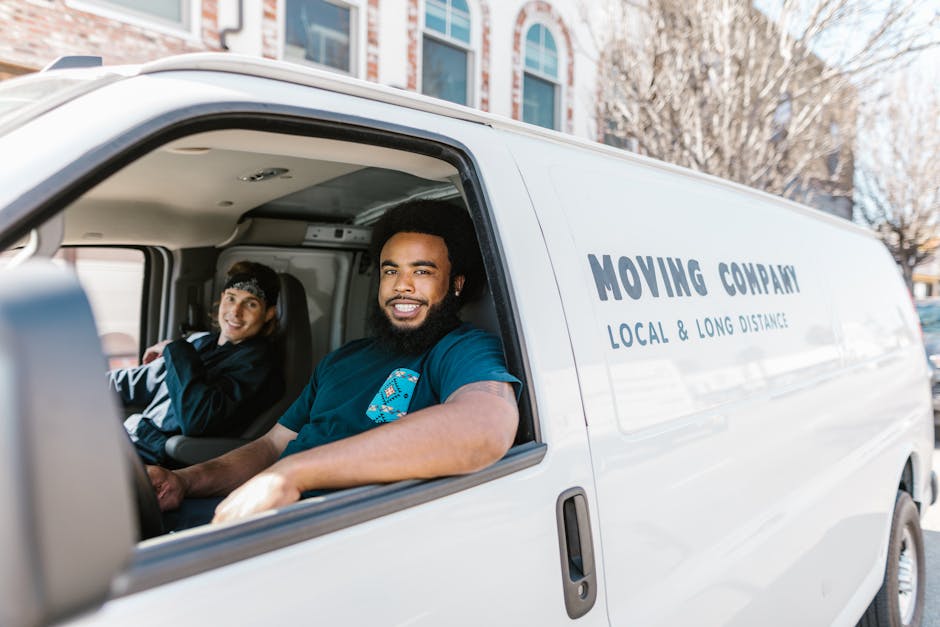 Two professional movers from Man with Van Millwall dressed in white protective suits, face masks, and blue gloves are loading cardboard boxes into the rear of a white moving van parked outdoors on a p