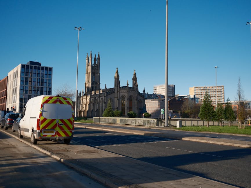 A white delivery van with red and yellow reflective chevron markings on the rear is parked on the side of a city street, adjacent to a sidewalk. The van is positioned facing away from the camera, and its rear doors are closed. In the background, there is a historic Gothic-style church with tall spires, surrounded by modern office buildings and residential apartments. The street is lined with small trees and lampposts, and a few cars are visible parked along the curb. The sky is clear with bright sunlight, casting shadows on the pavement. The scene depicts urban logistics involved in home relocation or furniture transport, possibly related to a professional removals service like [COMPANY_NAME], supporting packing and moving operations in Millwall or similar areas.