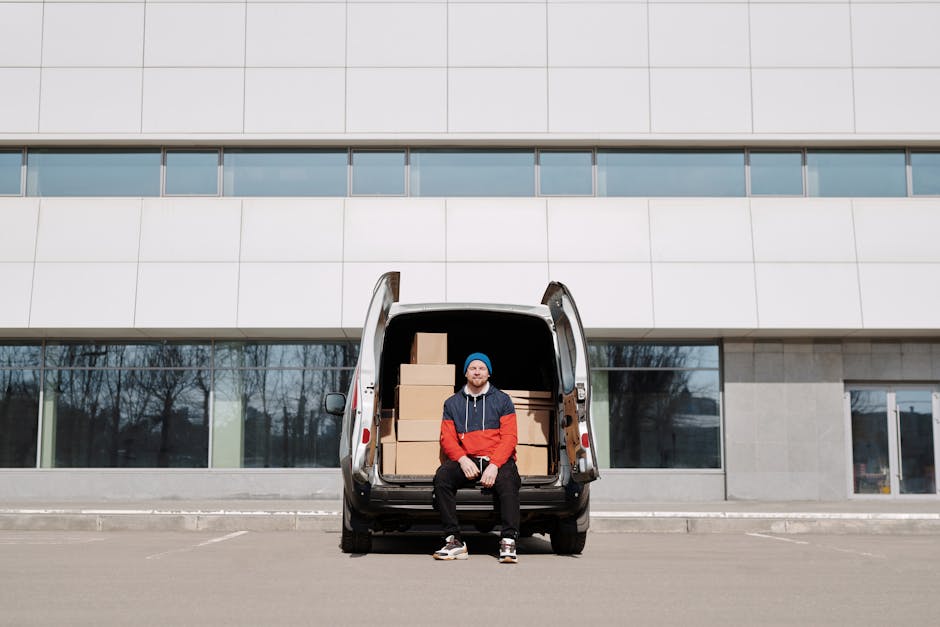 A man wearing a blue beanie, red and navy jacket, and black pants is seated on the edge of an open van's rear door in an outdoor parking area, with a modern multi-storey building with large windows in the background. The van's cargo area contains several cardboard boxes stacked neatly, secured for transport during a home relocation or furniture transport process. The scene shows the loading or unloading stage, with the van positioned on a paved surface and the boxes ready for transfer from or into a property. The image emphasizes professional packing, loading procedures, and transportation logistics involved in house removals, as offered by Man with Van Millwall.