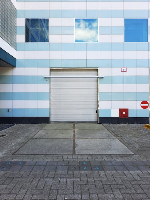 The image shows a modern commercial or industrial building with a light blue and white checkered façade, featuring four rectangular windows near the top, with shutters partially visible through the glass. Below the windows, there is a large, closed, white sectional roller shutter door, which is used for loading and unloading goods. To the right of the shutter, a red circular 'no entry' traffic sign with a white horizontal bar is mounted on the wall, and beneath it, a red rectangular fire safety box is also visible. The ground in front of the building comprises a spacious paved loading area with a gently inclined ramp leading up to the roller shutter, flanked by brick paving with a defined edge. The loading bay area appears clean and well-maintained, suitable for furniture transport and home relocation activities. This setup supports professional removals services by providing a clear, accessible space for loading and unloading items, often managed by companies like Man with Van Millwall during the packing and moving process near Millwall Dock.