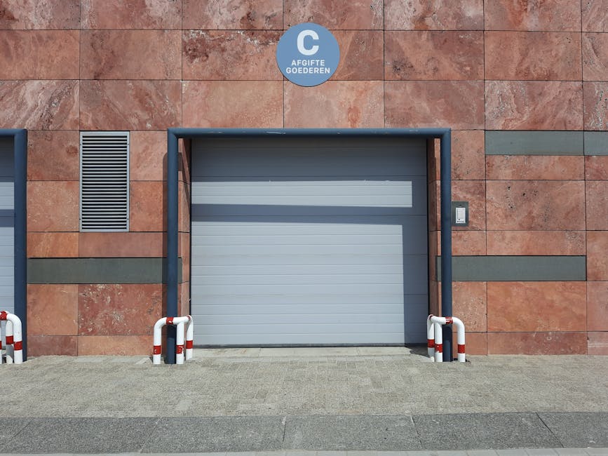 A grey, metal roller shutter garage door is situated within a brick wall exterior of a building, framed by a dark blue metal border. Above the garage, there is a circular sign with the text 'AFGIJTE GOEDEREN' in white letters on a blue background. To the left of the garage, there is a rectangular ventilation grille set into the wall. The ground in front of the garage is paved with light grey concrete tiles, and on either side of the door, there are white plastic safety barriers with red reflective stripes. The image was captured in daylight with clear weather, highlighting the building's facade, which features reddish-brown bricks with some horizontal greyish-green decorative bands, serving as part of the exterior environment for a home relocation or furniture transport process supervised by Man with Van Millwall, as indicated by the context.