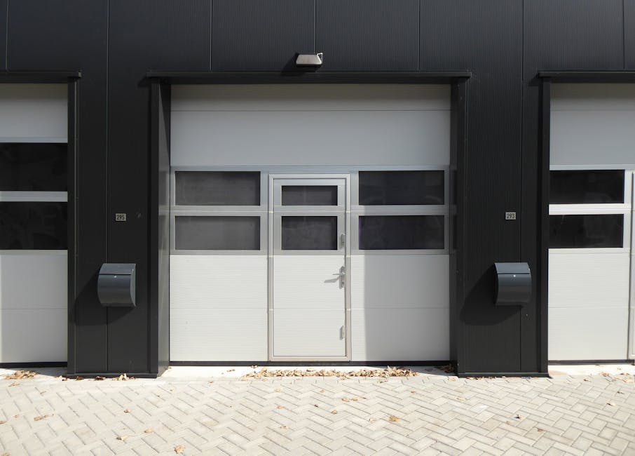 The image shows a modern commercial or industrial building with a light blue and white checkered façade, featuring four rectangular windows near the top, with shutters partially visible through the glass. Below the windows, there is a large, closed, white sectional roller shutter door, which is used for loading and unloading goods. To the right of the shutter, a red circular 'no entry' traffic sign with a white horizontal bar is mounted on the wall, and beneath it, a red rectangular fire safety box is also visible. The ground in front of the building comprises a spacious paved loading area with a gently inclined ramp leading up to the roller shutter, flanked by brick paving with a defined edge. The loading bay area appears clean and well-maintained, suitable for furniture transport and home relocation activities. This setup supports professional removals services by providing a clear, accessible space for loading and unloading items, often managed by companies like Man with Van Millwall during the packing and moving process near Millwall Dock.