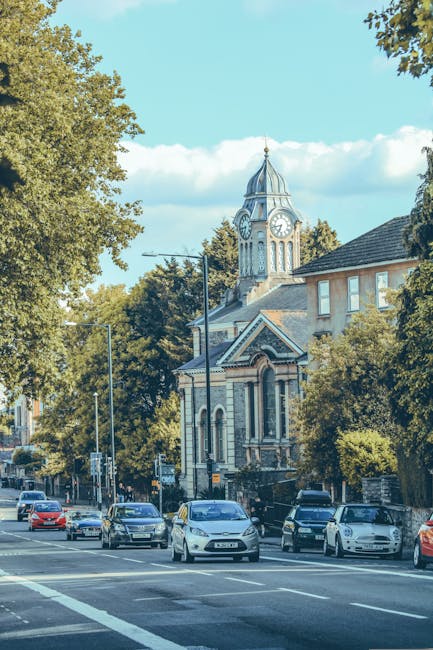 A row of parked cars along a street with a church featuring a tall clock tower and a domed spire visible in the background. The street is lined with large leafy trees, some of which cast shadows on the pavement. In the foreground, a temporary parking restriction sign is mounted on a pole. The scene is set during daytime with a clear sky and scattered clouds, indicating calm weather. The setting reflects an urban area suitable for home relocation and furniture transport services, illustrating the typical environment where Man with Van Millwall may operate, including the need for parking permits and efficient moving logistics within the town.