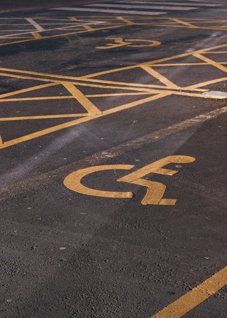 A close-up view of a parking area with freshly painted yellow wheelchair accessible parking symbols on dark asphalt surface. The parking bays are outlined with yellow lines, some of which are partially visible in the background. The area appears to be outdoors, with the pavement showing minor wear and some white marking lines for directional guidance. This image is relevant to house removals and moving services, highlighting accessible parking options that might be used during a home relocation. Man with Van Millwall offers professional removals and transport services, supporting clients with moves that require accessible parking permits from Tower Hamlets Council. The scene emphasizes the importance of designated parking spaces for safe and efficient furniture transport and loading processes during house moves.