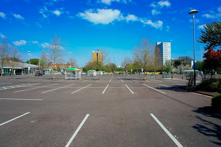 A row of parked cars along a street with a church featuring a tall clock tower and a domed spire visible in the background. The street is lined with large leafy trees, some of which cast shadows on the pavement. In the foreground, a temporary parking restriction sign is mounted on a pole. The scene is set during daytime with a clear sky and scattered clouds, indicating calm weather. The setting reflects an urban area suitable for home relocation and furniture transport services, illustrating the typical environment where Man with Van Millwall may operate, including the need for parking permits and efficient moving logistics within the town.