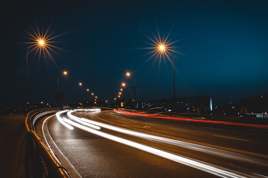 Long-exposure photograph of a curved city street at night, illuminated by tall streetlights emitting a warm, yellow glow that creates starburst effects. The road features white and red light trails from moving vehicles, indicating active traffic flow. On the left side, a guardrail runs parallel to the road, which appears to be part of a bridge or elevated roadway. In the background, there are dark silhouettes of buildings and infrastructure under a deep blue night sky. The scene captures the dynamic movement and urban environment, relevant for house removals and moving logistics services offered by Man with Van Millwall, especially emphasizing efficient transportation and route navigation in areas like Westferry Road, Millwall.