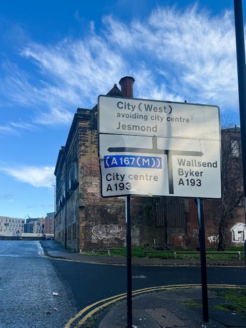 A roadside traffic sign mounted on two metal posts displays directional information for drivers on a city street. The sign indicates routes to the city centre, Jesmond, Wallsend, and Byker via the A193 and A167(M). The sign includes additional details such as avoiding the city centre and mentions Jesmond. Behind the sign, an old brick building with graffiti on its lower walls and a chimney on the roof is visible, along with a paved road that curves to the left and features yellow double lines along the curb. The sky above is partly cloudy with patches of blue, and the overall environment suggests an urban setting. This scene relates to house removals and moving logistics, emphasizing navigation routes for efficient transport planning, as part of professional services like those offered by Man with Van Millwall.