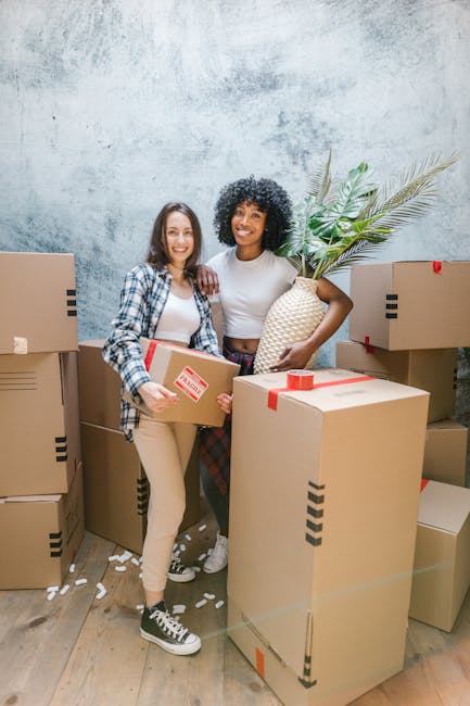Two women engaged in a home relocation process indoors, surrounded by stacked cardboard boxes sealed with red tape, some labeled with handling instructions. The women are standing close together, smil