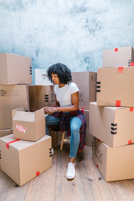 Two women engaged in a home relocation process indoors, surrounded by stacked cardboard boxes sealed with red tape, some labeled with handling instructions. The women are standing close together, smil