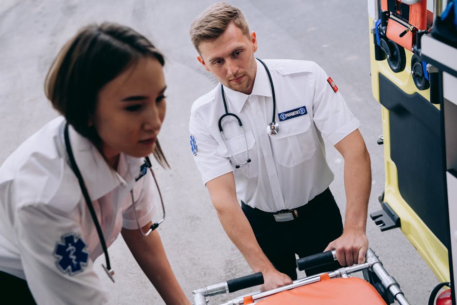 Two healthcare professionals, a female and a male paramedic, are preparing medical equipment for transportation. They are dressed in white uniforms with stethoscopes around their necks, and their focus is on handling a wheeled stretcher with an orange sheet covering the padded surface. The female paramedic is slightly out of focus in the foreground, while the male paramedic is more centered, gripping the handles of the stretcher. Behind them, part of an emergency vehicle with open compartments containing medical supplies and equipment is visible. The scene takes place indoors, likely in a hospital or ambulance station, with a concrete floor and neutral lighting. This image illustrates the process of emergency medical response and patient transport, emphasizing professional care and readiness, consistent with the themes of medical and patient relocation services.