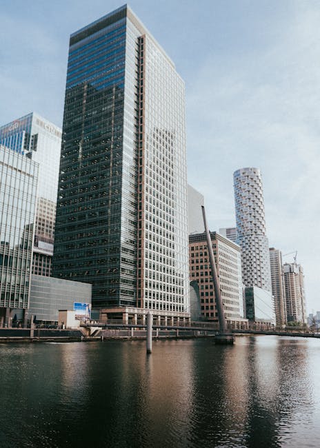 A daytime view of a modern city skyline featuring several high-rise office buildings with glass facades reflecting the sky and surrounding structures, situated along a river with calm water. The tallest building has a rectangular shape with a grid-like window pattern. Nearby, there are other tall buildings with various architectural styles, including rounded and angular designs, some under construction with cranes visible. In the foreground, a concrete walkway runs alongside the water, and a metal railing is visible along the edge. This urban scene exemplifies a busy business district near the river, relevant to house and office relocations, with the potential involvement of professional removals like those by Man with Van Millwall for logistics planning and furniture transport in areas such as Millwall and Canary Wharf.