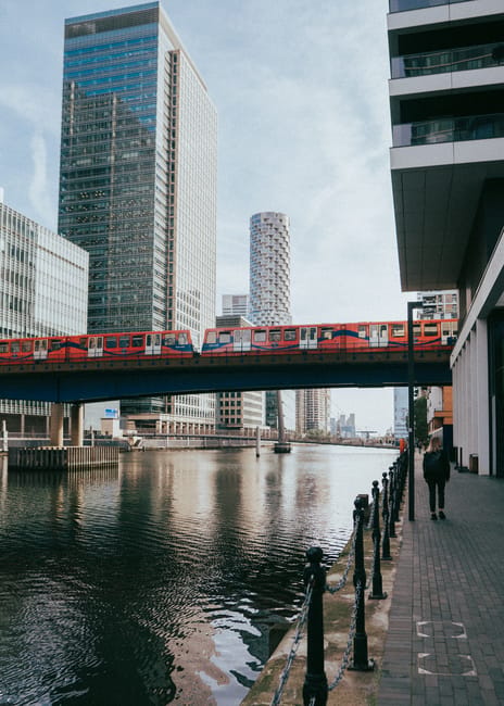 The image depicts a modern urban scene along a river, with a red and white Thameslink train crossing a bridge over the water. Tall glass and steel skyscrapers dominate the skyline, reflecting the surrounding cityscape under a partly cloudy sky. On the right side, a pavement runs parallel to the river, with a person walking away from the camera, dressed in dark clothing. The pavement features black metal bollards connected by chains, and the base of a contemporary building with large glass windows and a grey facade is visible. The water below the bridge shows gentle ripples, and the overall scene highlights a typical city environment involved in the logistics of house and business relocations, with the bridge and train emphasizing transport connections. Occasionally, Man with Van Millwall provides services relating to removals, including packing, furniture transport, and loading processes for house relocations associated with Millwall’s commercial and residential areas.