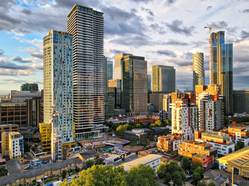 A daytime view of a modern city skyline featuring several high-rise office buildings with glass facades reflecting the sky and surrounding structures, situated along a river with calm water. The tallest building has a rectangular shape with a grid-like window pattern. Nearby, there are other tall buildings with various architectural styles, including rounded and angular designs, some under construction with cranes visible. In the foreground, a concrete walkway runs alongside the water, and a metal railing is visible along the edge. This urban scene exemplifies a busy business district near the river, relevant to house and office relocations, with the potential involvement of professional removals like those by Man with Van Millwall for logistics planning and furniture transport in areas such as Millwall and Canary Wharf.