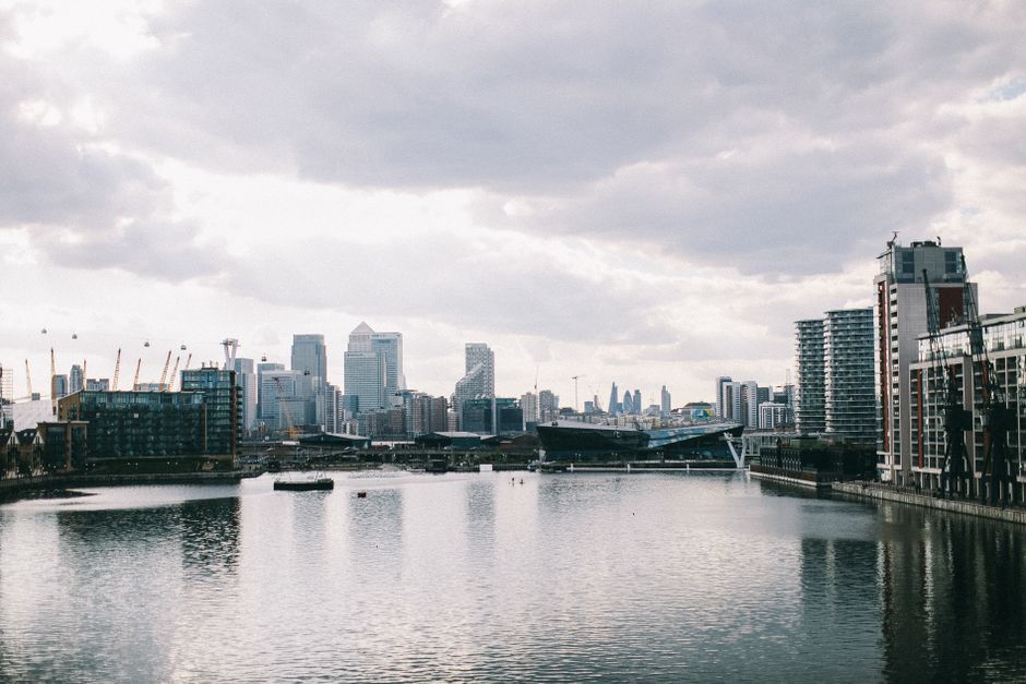 A wide view of the London skyline overlooking the River Thames during daytime, with modern high-rise buildings, cranes in the background, and partially cloudy skies. In the foreground, calm water reflects the surrounding structures, and along the riverbank to the right, there are residential and commercial buildings with glass facades. This scene captures an urban setting relevant to house removals and logistical planning for property relocation, illustrating the proximity of city infrastructure to residential areas. The image emphasizes city-based moving services such as furniture transport and home relocation, aligning with the services offered by Man with Van Millwall, especially for residential moves within or into the Canary Wharf and Millwall areas.