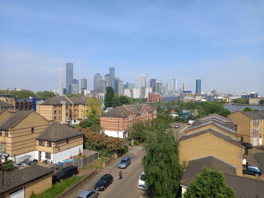 The image captures a daytime view including a row of residential houses with brick and beige exteriors, sloped tiled roofs, and small front yards with trees and parked cars along a narrow street. In the background, a modern city skyline featuring numerous high-rise office buildings with glass facades and varied architectural styles is visible under a clear blue sky. The foreground shows the residential area, likely part of a home relocation or furniture transport process, with a focus on the urban environment. The scene suggests a peaceful neighbourhood adjacent to a busy commercial district, demonstrating the typical context for professional removals and moving services such as those offered by Man with Van Millwall.