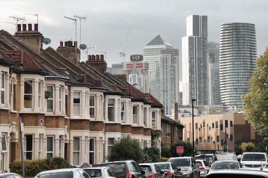 A wide view of the London skyline overlooking the River Thames during daytime, with modern high-rise buildings, cranes in the background, and partially cloudy skies. In the foreground, calm water reflects the surrounding structures, and along the riverbank to the right, there are residential and commercial buildings with glass facades. This scene captures an urban setting relevant to house removals and logistical planning for property relocation, illustrating the proximity of city infrastructure to residential areas. The image emphasizes city-based moving services such as furniture transport and home relocation, aligning with the services offered by Man with Van Millwall, especially for residential moves within or into the Canary Wharf and Millwall areas.